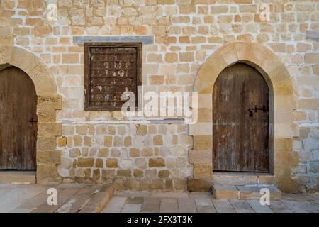 Due porte intemperate in legno ad arco e una finestra chiusa in ferro battuto arrugginito su mattoni parete in pietra e pavimento in piastrelle di pietra Foto Stock