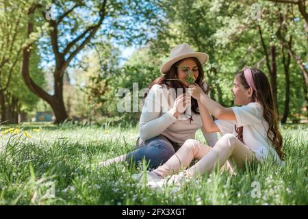 Madre che annuisce i fiori che sua figlia ha raccolto nel parco estivo. La famiglia si diverte all'aperto rilassandosi sull'erba Foto Stock