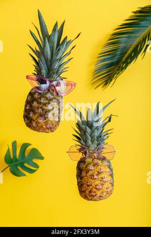 Ananas facce creative in occhiali da sole a colori con foglie di palma su sfondo estivo di colore giallo. Frutti tropicali estivi che levitano ananas. Foto Stock