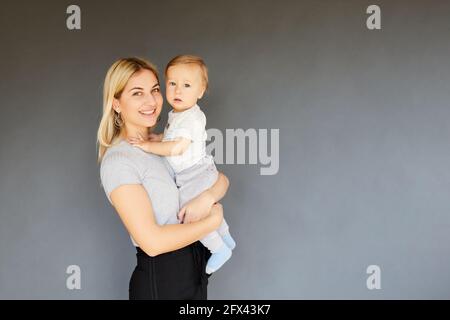 Sorridente bella donna che tiene il suo bambino di undici mesi con sfondo grigio. Concetto di maternità Foto Stock