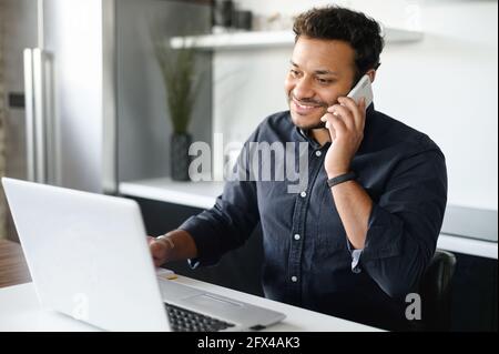Un ragazzo indiano parla sullo smartphone e utilizzando un computer portatile che si trova a casa e lavora a distanza, sorridendo uomo indù ha conversazione telefonica guardando lo schermo del computer Foto Stock