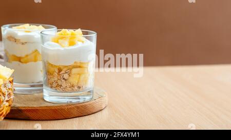 Gustosi dessert di ananas con sminuzzati di ananas fresco e succoso. Prima colazione dessert a base di granola d'avena, yogurt greco e strati di ananas in vetro su tavolo di legno Foto Stock