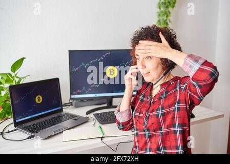 Scioccata donna trader mette le mani alla sua testa a causa al crollo del mercato della criptovaluta mentre si parla telefono cellulare Foto Stock