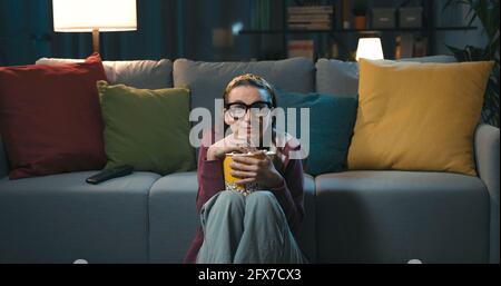 Donna allegra guardando la sua serie preferita in televisione, è seduta sul pavimento e mangiare popcorn Foto Stock