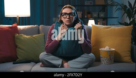 Donna spaventata seduta sul divano e guardando un film horror in TV, lei sta abbracciando un cuscino Foto Stock