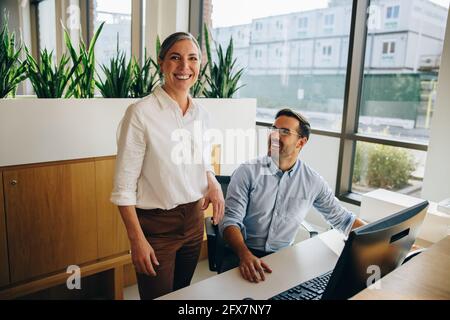 Donna matura in piedi e sorridente alla macchina fotografica mentre il giovane uomo si siede alla scrivania. Felici i colleghi in ufficio al lavoro. Foto Stock
