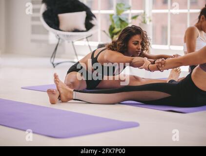 Due giovani donne che si allenano in coppia al chiuso. Amici seduti sul tappeto yoga che tengono le mani e si allungano insieme. Foto Stock