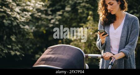 Mamma che cammina intorno al quartiere spingendo il suo bambino nel passeggino. Donna che controlla i messaggi sul suo smartphone mentre spinge un pRAM. Foto Stock