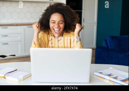 Eccitata felice afro-americana freelance donna gridare sì, guardando lo schermo del portatile, celebrando la vittoria, obiettivo raggiunto, nero femmina con acconciatura sollevata pugni in su, ha ricevuto opportunità Foto Stock
