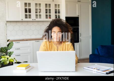 Affascinante ragazza africana curly che lavora con un computer portatile da casa. Donna freelance intelligente con pelle scura che utilizza il computer per lavorare a distanza, digitando sulla tastiera, inviando e-mail. Studentessa online Foto Stock