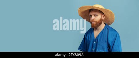 Giovane uomo rosso con testa e bearded in cappello di paglia isolato su bacground blu. Volantino Foto Stock