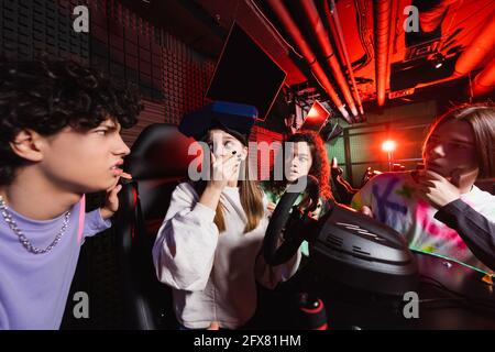 scioccata ragazza su simulatore di corsa che copre la bocca con la mano vicino amici interrazziali Foto Stock