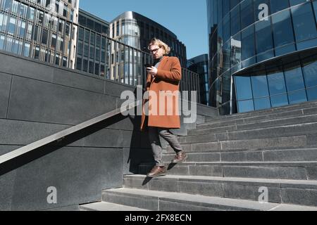 Giovane uomo che digita un messaggio sul telefono cellulare mentre si è in movimento giù per le scale in città Foto Stock