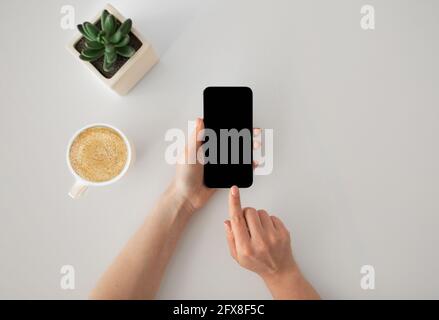 Concetto di pausa caffè. Smartphone da donna a contatto con la mano con schermo nero vuoto, seduto al tavolo, vista dall'alto Foto Stock