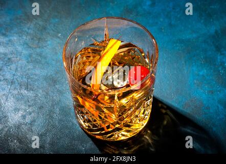 Cocktail alcolico vecchio stile con whisky di bourbon, ciliegia di maraschino e contorno di buccia d'arancia, tavolo blu, spazio per la copia, vista dall'alto, luce dura Foto Stock