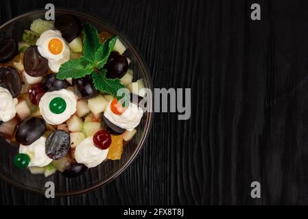 Insalata di uva mele pere arance kiwi con mascarpone chease e crema. Insalata estiva di frutta fresca sana in una ciotola di vetro su un backgroun di legno nero Foto Stock