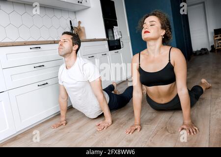 Concentrata coppia facendo esercizi di yoga sul pavimento della cucina moderna insieme, facendo cane faccia in su pone allenamento indossando abbigliamento sportivo, indoor pieno Foto Stock