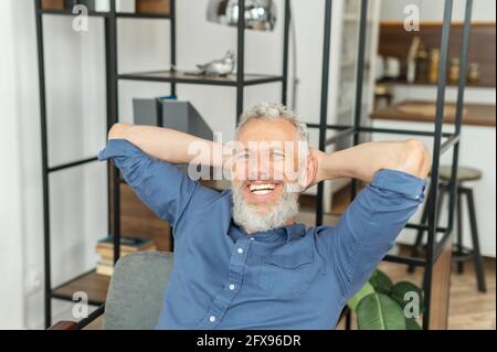 Rilassato allegro uomo dai capelli grigi senior riposante in ufficio, uomo d'affari carismatico maturo in camicia casual elegante godendo di finitura di buona giornata di lavoro, giorno di partenza, tenendo le mani dietro la testa Foto Stock