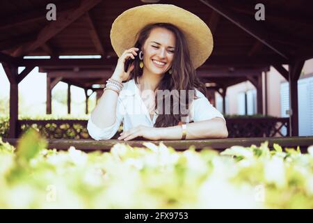 sorridente donna trendy in camicia bianca con cappello utilizzando uno smartphone sul ranch. Foto Stock