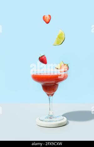 Ingredienti che cadono al cocktail Margarita Strawberry su sfondo blu. Orientamento verticale. Bevanda alcolica estiva fresca. Foto Stock