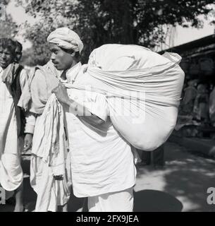 Anni '50, storico, fuori in una zona di mercato di una città, un giovane indiano, uno Skih, indossando un turbante di stoffa, forse un keski, che trasporta un carico grande, una coperta grande riempita di vestiti e lenzuola, India. Foto Stock