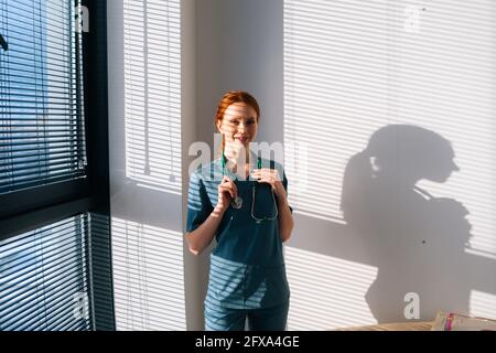 Ritratto di felice dottore in uniforme verde blu in piedi vicino alla finestra in giornata di sole in clinica medica ufficio. Foto Stock