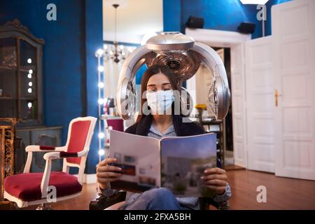 Una donna che legge una rivista con la sua maschera in un salone di bellezza Foto Stock