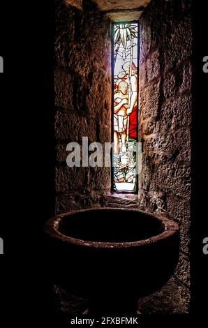 Fonte battesimale in una chiesa medievale illuminata da una macchiata finestra di vetro con scene bibliche Foto Stock