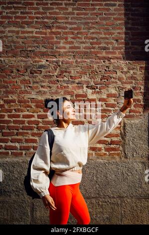 Sportswoman con zaino che porta i selfie attraverso lo smartphone mentre si trova di fronte alla parete in mattoni Foto Stock