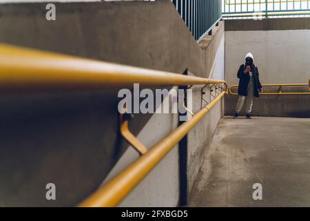 Donna che indossa una giacca con cappuccio e una maschera protettiva per il viso utilizzando il telefono cellulare in sottosuolo Foto Stock