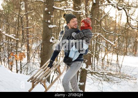 Padre con la slitta che tiene il figlio mentre cammina nella neve durante l'inverno Foto Stock