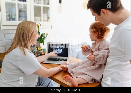 Donna incinta sorridente che mostra l'immagine ecografica alla figlia da padre sul tavolo in soggiorno Foto Stock