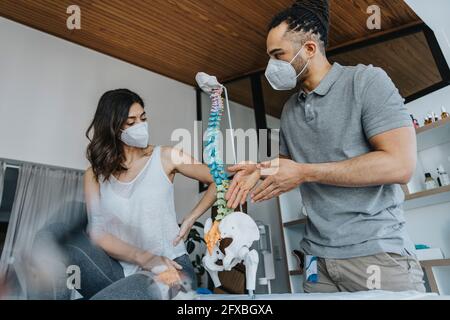 Fisioterapista maschile gesturing mentre discute con paziente femminile sopra modello di spin nella pratica durante COVID-19 Foto Stock