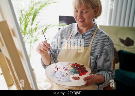 Donna sorridente che mescola i colori mentre fa la pittura a casa Foto Stock