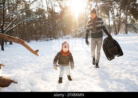 Padre con slitta che cammina verso il figlio sulla neve durante l'inverno Foto Stock