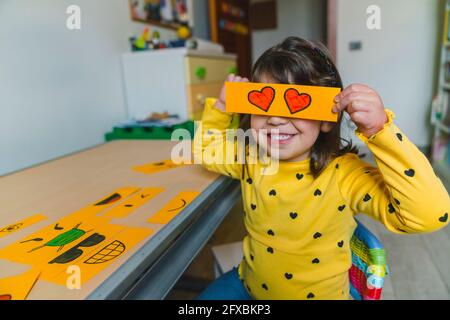 Ragazza che tiene il cuore forma emoticon sopra gli occhi mentre gioca a casa Foto Stock