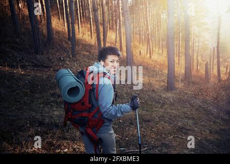 Donna sorridente con zaino che guarda sopra la spalla mentre si fa un'escursione in bosco Foto Stock