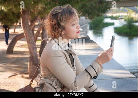 Bella ed elegante donna di mezza età che fa un selfie con lei telefono cellulare in un parco appoggiato alla parete la riva di un fiume in città Foto Stock