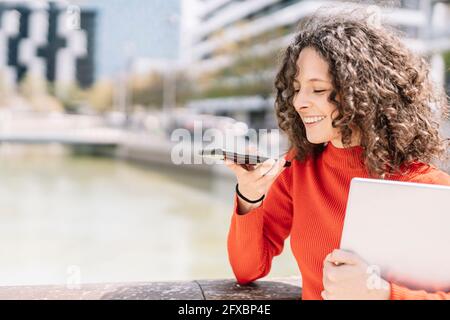 Donna che invia messaggi vocali tramite telefono cellulare mentre tiene il computer portatile con la ringhiera Foto Stock