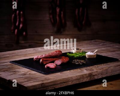 Salsiccia di Landjager con rosmarino, sale e pepe in ciotole e semi di girasole su lastra di pietra ardesia nera Foto Stock
