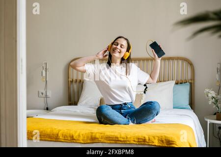 Donna allegra con telefono cellulare che ascolta la musica attraverso le cuffie in camera da letto Foto Stock