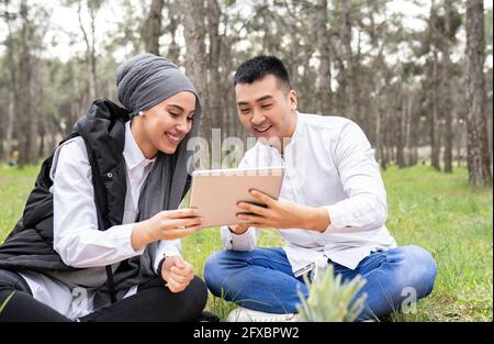 Sorridente giovane e bella donna che usa il tablet digitale mentre si siede in foresta Foto Stock