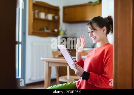 Donna sorridente che sventolava la mano per videochiamare sul tablet digitale a casa Foto Stock