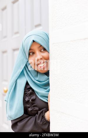 Donna sorridente che indossa hijab sbirciando intorno al muro Foto Stock