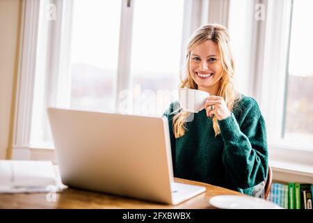 Donna d'affari sorridente seduto con il computer portatile che beve caffè in ufficio Foto Stock