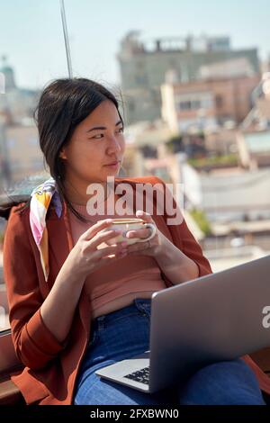 Donna premurosa seduta con computer portatile e tazza di caffè sulla terrazza sul tetto Foto Stock