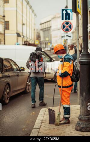 Ragazza lavoratore pulitore in tute luminose in maschera monouso viso con strumento scopa e smartphone sulla strada della città Foto Stock