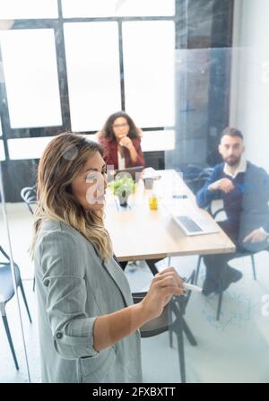 Donna d'affari che discute il programma di affari con i colleghi di maschio e femmina visti attraverso il vetro all'ufficio Foto Stock