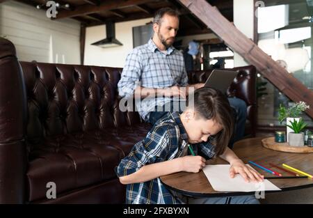 Ragazzo che disegnava su carta mentre padre usa il computer portatile sul divano a casa Foto Stock