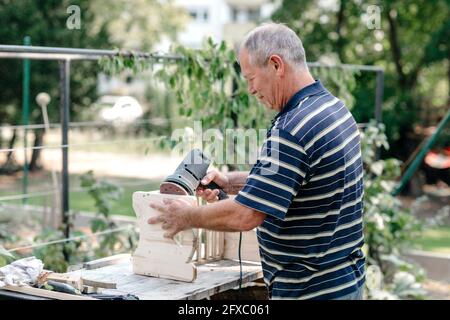 Uomo anziano attivo con l'attrezzo di lavorazione del legno che fa la carpenteria in cortile Foto Stock